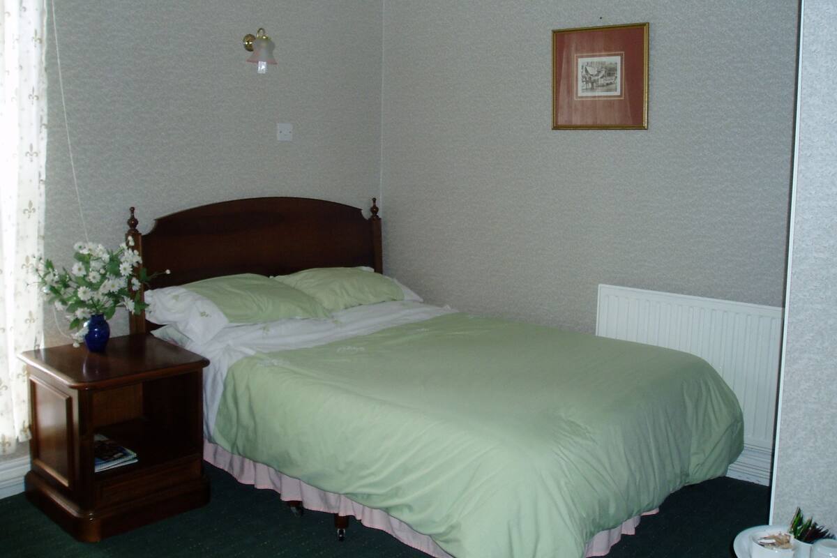 Un lit bien fait, recouvert d’une couverture verte et agrémenté d’oreillers blancs, une table de chevet en bois avec un vase de fleurs, et un tableau encadré accroché au mur.