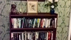 A bookshelf with books and decorative items, a framed picture above it, and a patterned rug on the floor. - Rhymney House hotel (Tredegar)