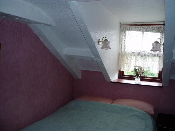 A bedroom with a sloped ceiling, a window with lace curtains, a bed with pillows, and a nightstand.