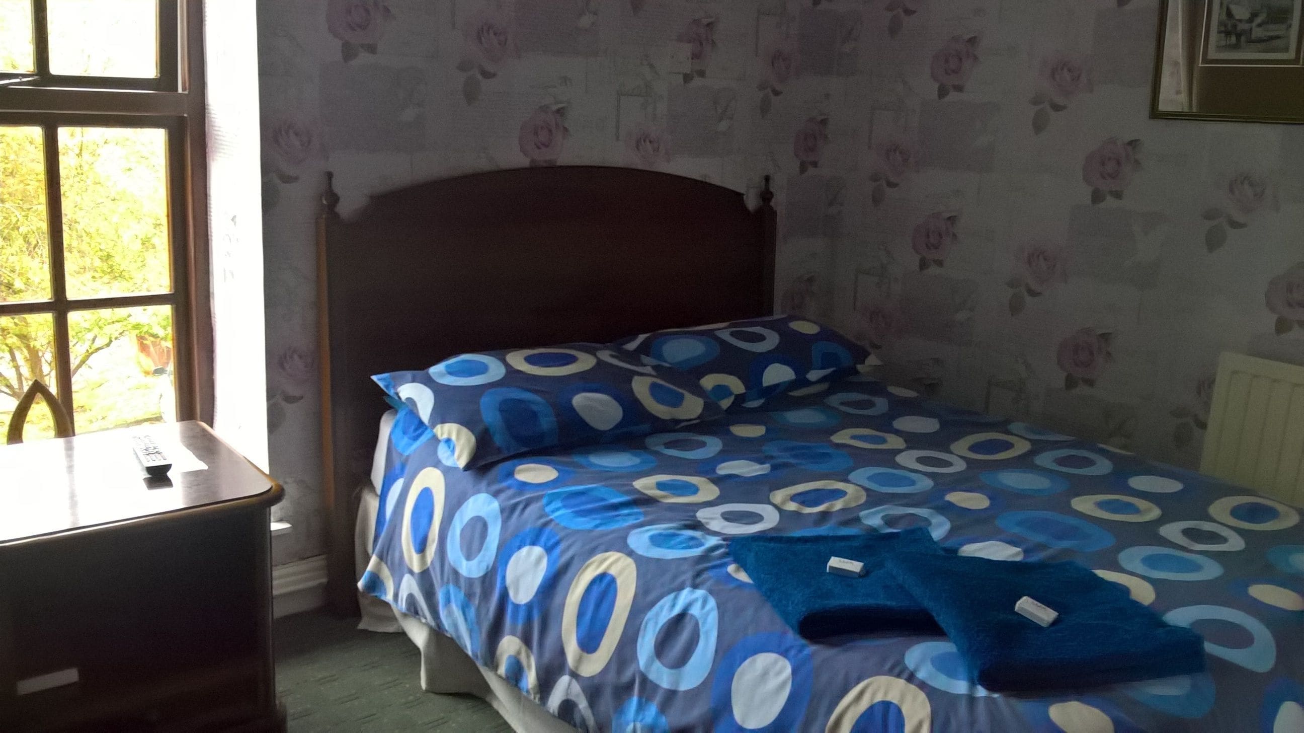 A bedroom with a bed, a nightstand, a window, and a floral wallpaper.