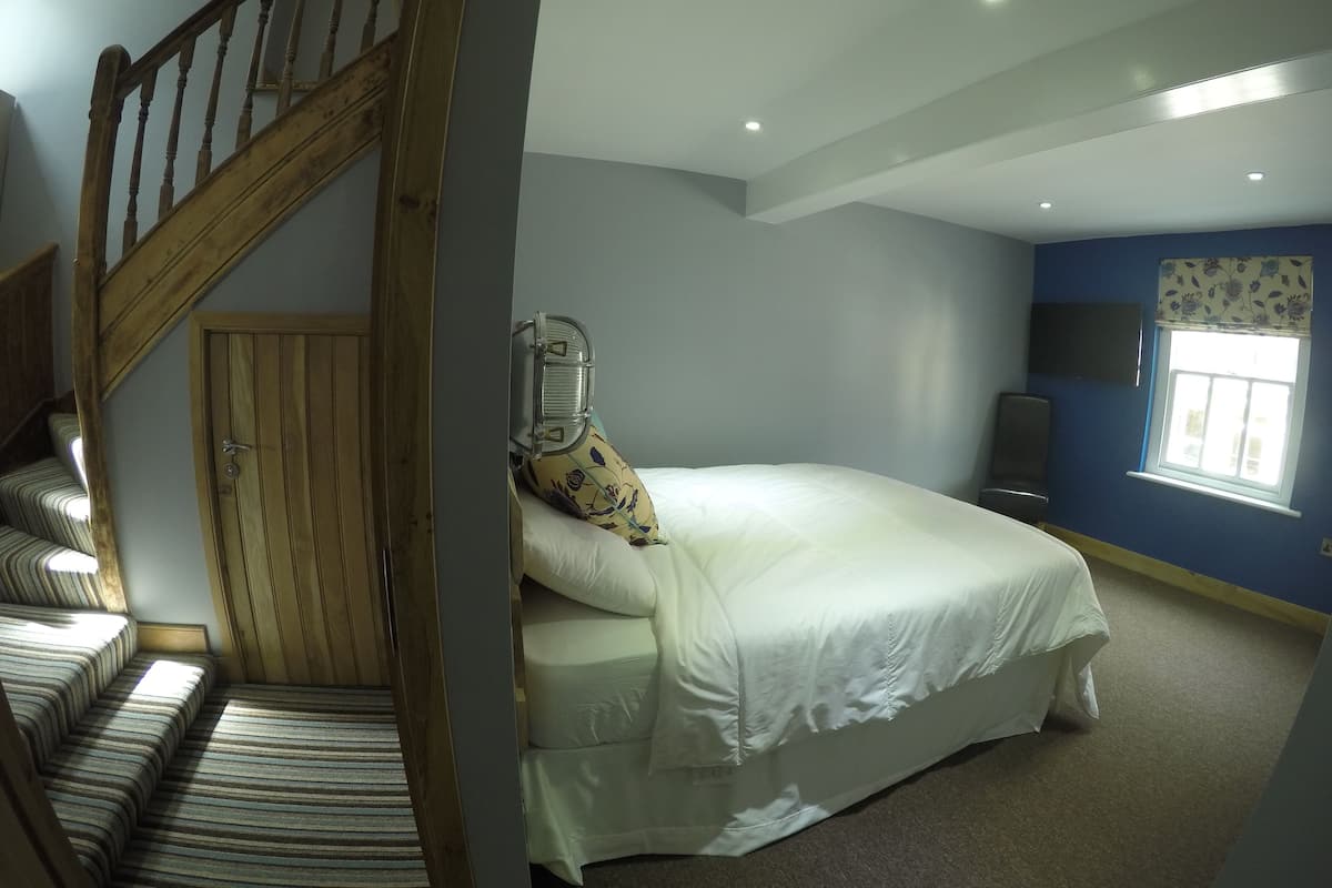 A bedroom with a bed, a wooden staircase, a window with blinds, and a flat-screen TV on the wall.