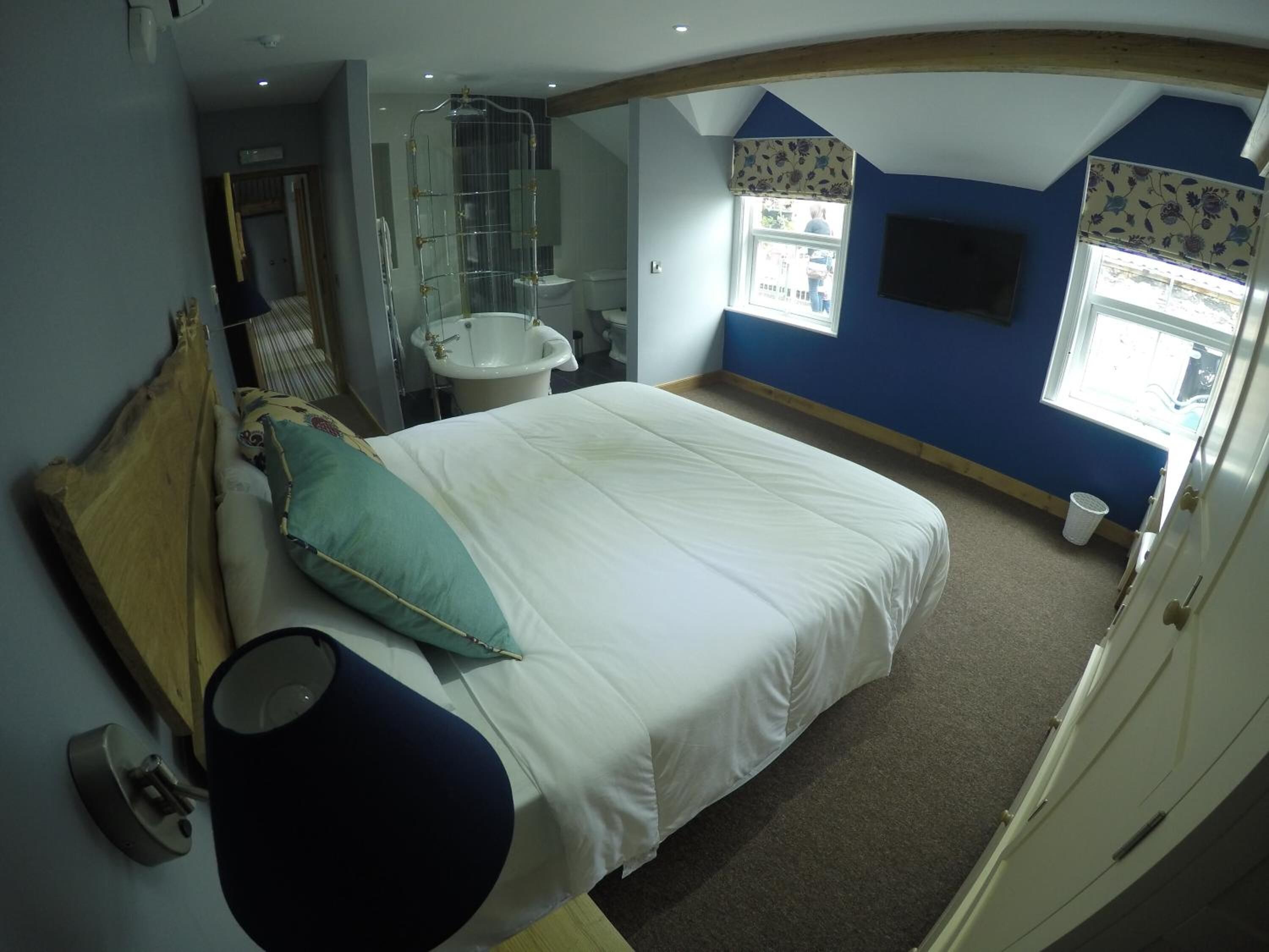 A bedroom with a bed, a bedside table, a mirror, a window, and a bathroom with a sink and shower.