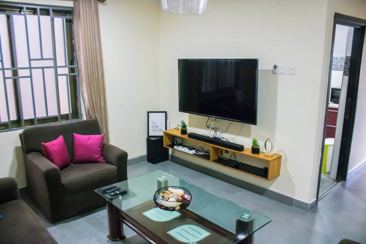 Una sala de estar con un sofá, una mesa de centro de vidrio, un televisor de pantalla plana y una estantería de madera con adornos.