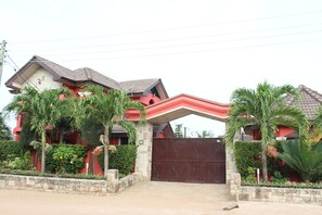 A two-story house with a red facade, a gated entrance, and a small garden with palm trees. - Nego Lodge (Accra)