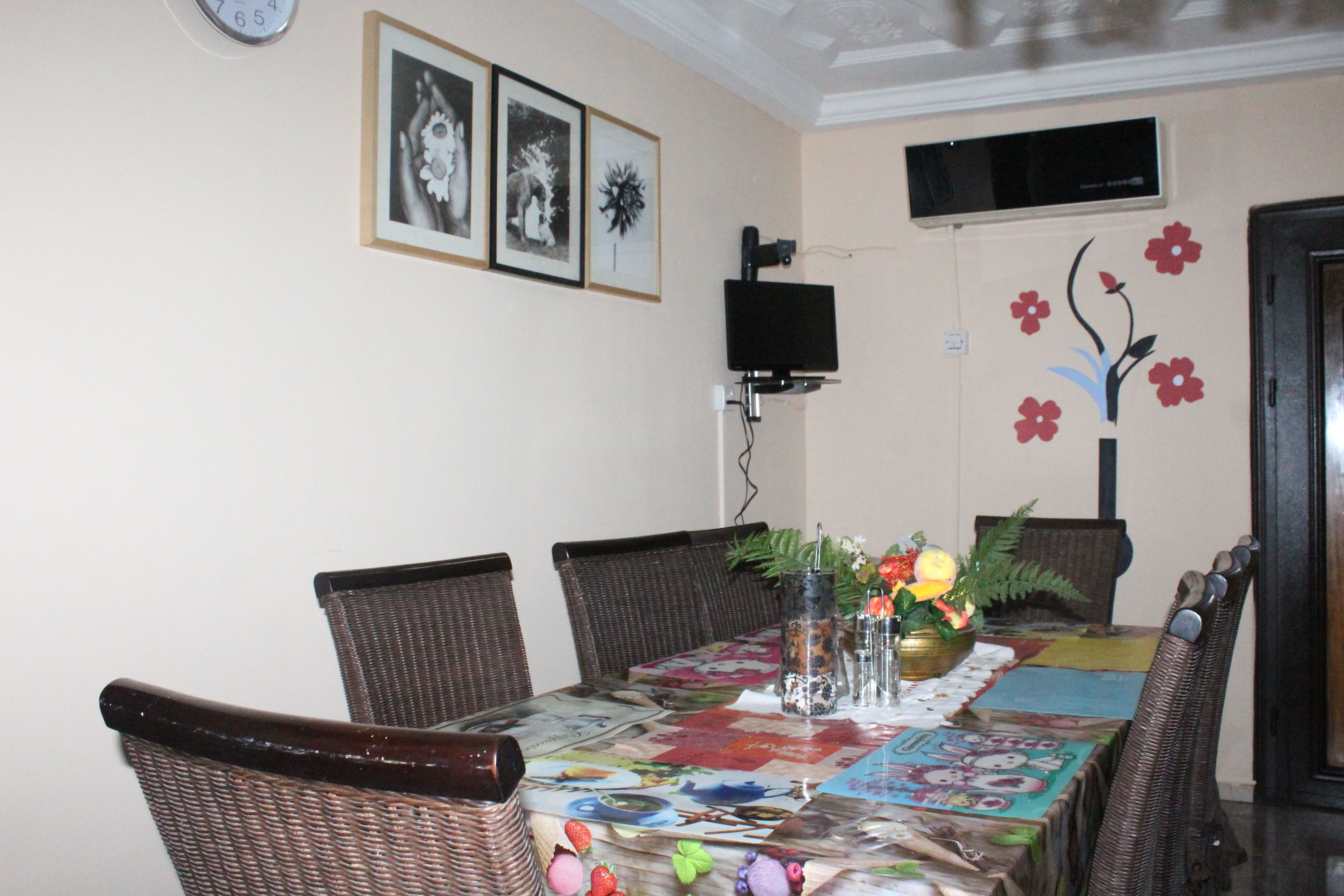 A dining table with a floral tablecloth, surrounded by wicker chairs, a clock on the wall, and a television mounted above.