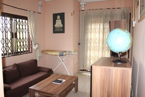 A living room with a brown sofa, a wooden coffee table, a globe lamp, a wall-mounted Buddha statue, and a window with curtains. - Nego Lodge (Accra)