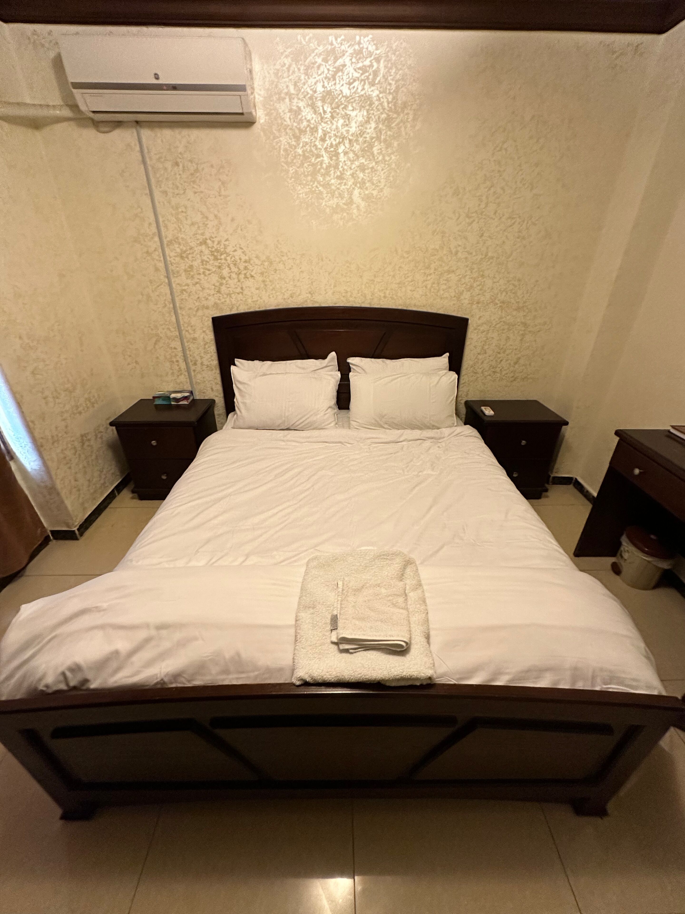 A neatly made bed with white linens and a folded towel, two bedside tables with lamps, and an air conditioning unit on the wall.