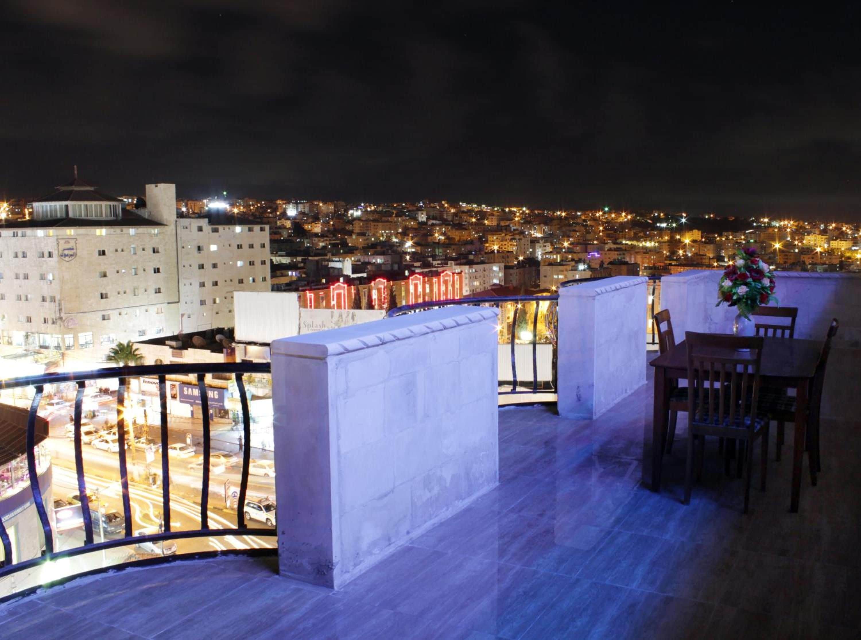 A rooftop terrace with a view of a cityscape at night, featuring a dining area with chairs and a table, and a flower arrangement.