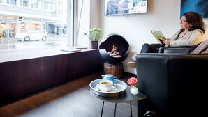 Un salon moderne doté d’une cheminée, d’une table basse sur laquelle se trouve une tasse de café et une rose, ainsi qu’un téléviseur diffusant des images d’un groupe de personnes.