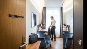 Une chambre d’hôtel avec une porte en bois portant l’inscription « RENNWEG », un bureau, deux chaises, un téléviseur et une grande fenêtre avec des rideaux.