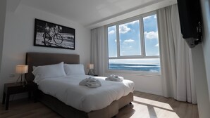 A hotel room with a large bed, a view of the sea, a painting on the wall, and a TV mounted on the wall.