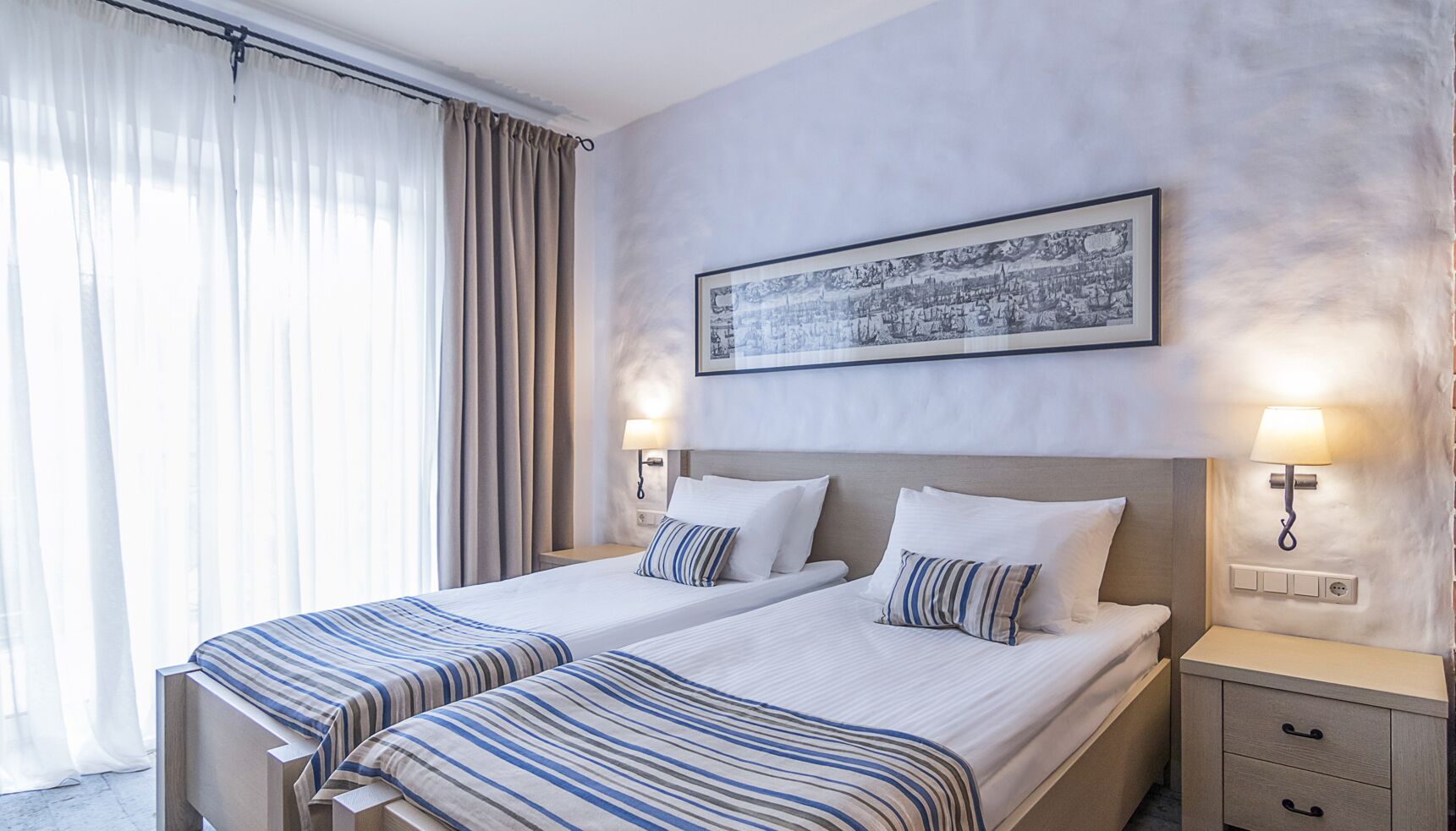 A hotel room with two beds, a large window with curtains, a framed picture on the wall, and bedside lamps.