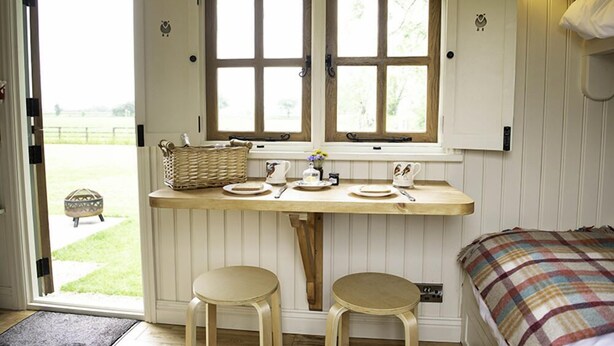 A cozy room with a wooden table, two stools, a bed with a plaid blanket, and a window with a view of the outdoors. - Morndyke Shepherds' Huts (Thirsk)