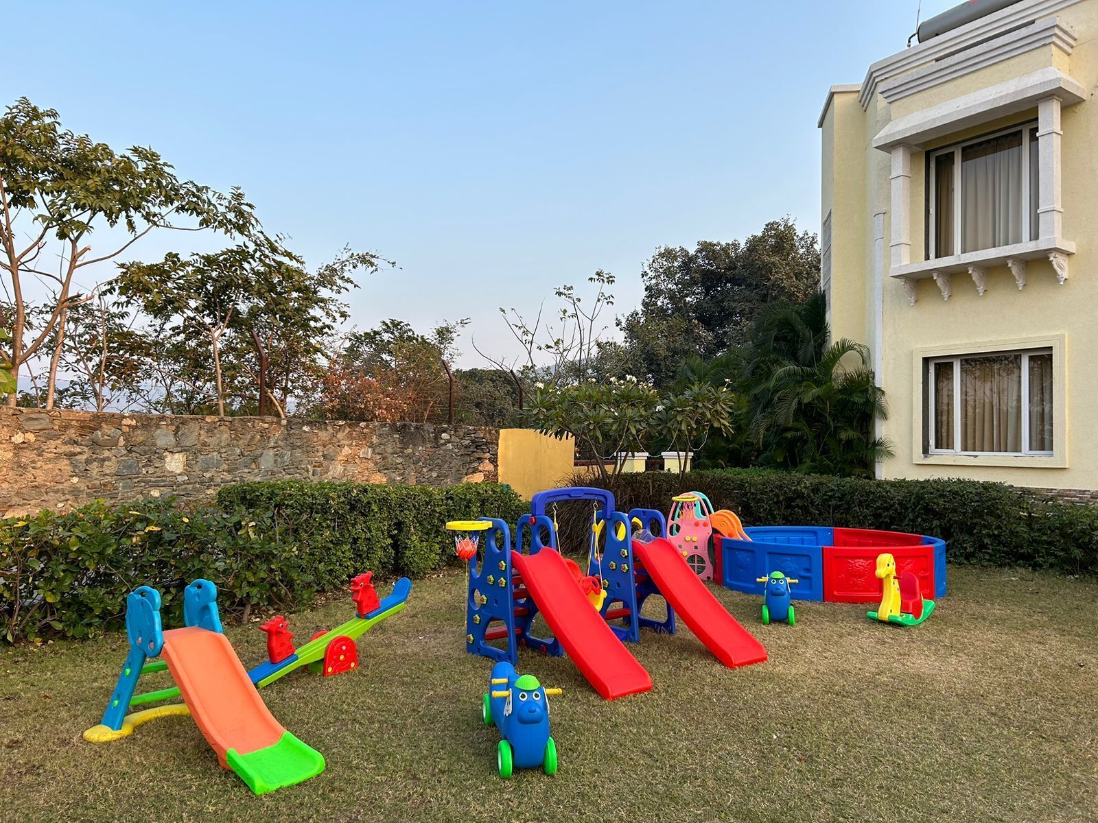 A playground with various play structures including slides and climbing frames, surrounded by a stone wall and greenery.