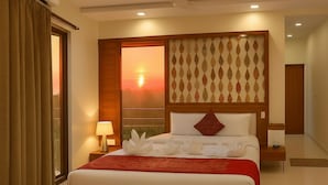 A hotel room with a large bed, a nightstand with a lamp, a window with a sunset view, and a ceiling fan. - Alcor Kumbhalgarh – the family getaway (Kumbhalgarh)
