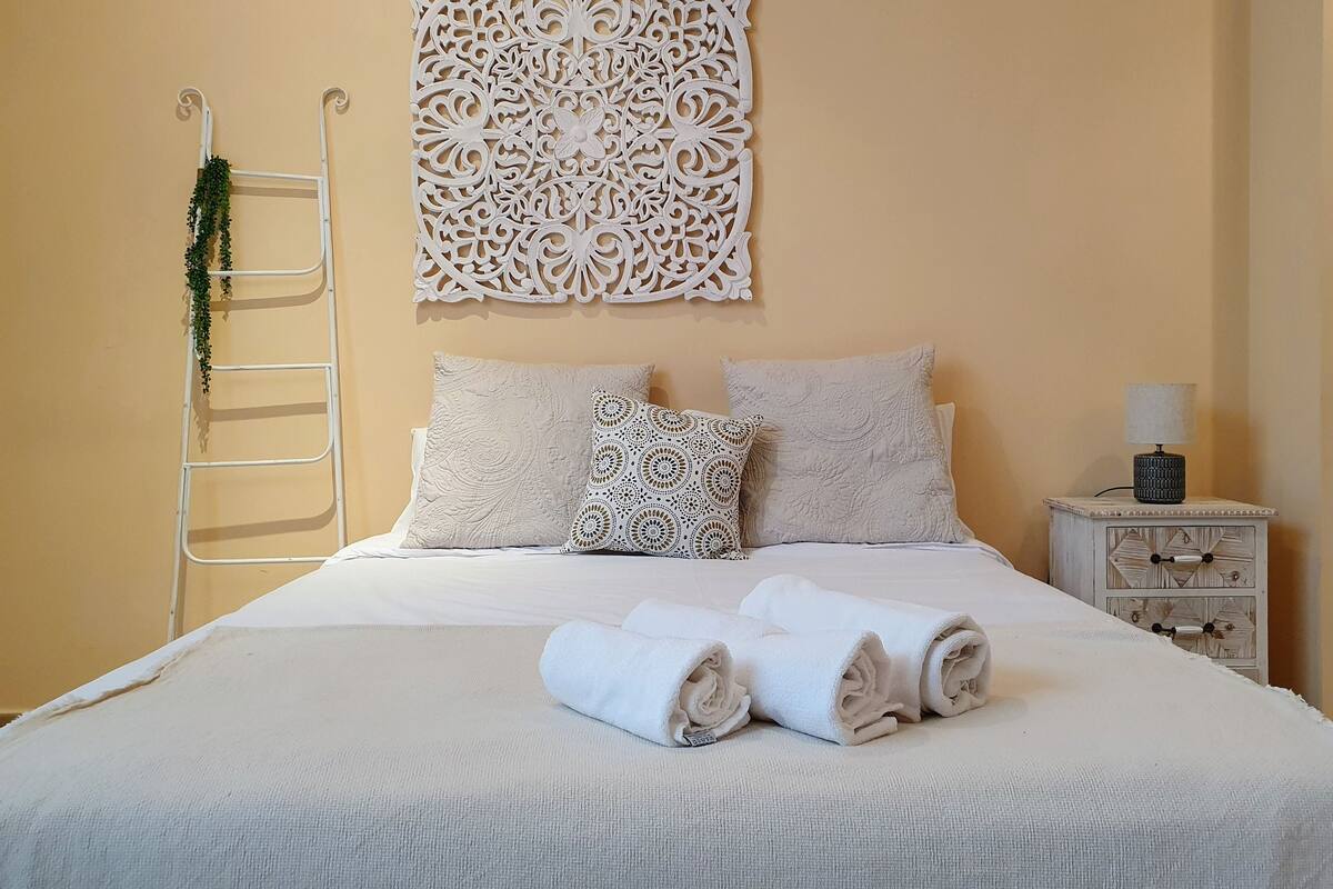 A neatly made bed with rolled towels, a decorative wall piece, a bedside table with a lamp, and a white metal ladder with greenery.