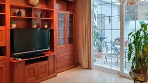 Un salón con televisor sobre un mueble de madera, una araña y vistas al patio exterior.