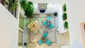 Un patio interior con una mesa redonda central rodeada de cuatro sillas, una planta grande en maceta y un adorno en el techo.