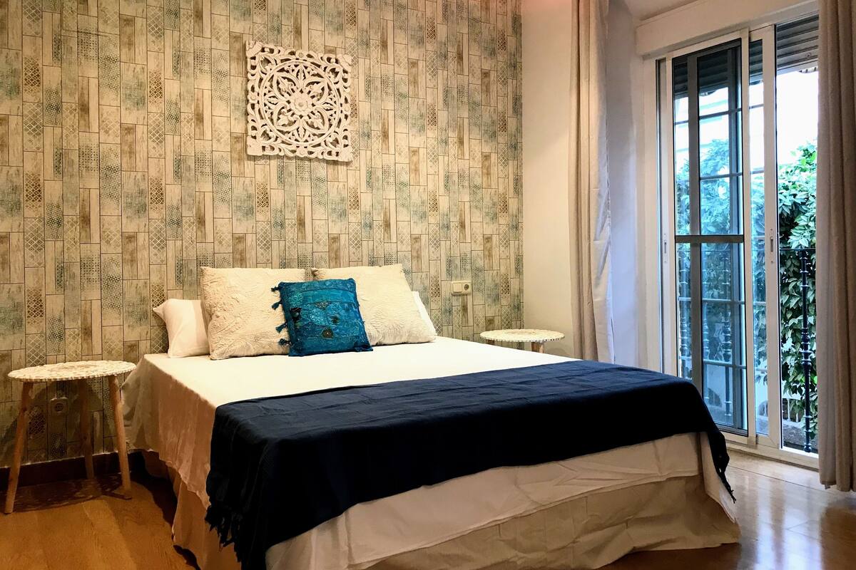 A bedroom with a patterned wallpaper, a bed with a black and white bedspread, two bedside tables, and a window with curtains.