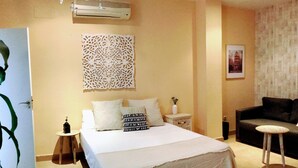 Habitación de hotel con una cama grande, una mesita auxiliar pequeña, un sofá, una araña y un elemento decorativo en la pared.