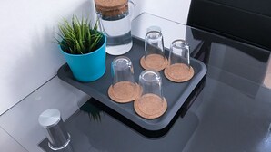 A glass table with a cork coaster tray holding a glass jar with a cork lid, a blue pot with green plants, and several clear glass bottles. - Charles Bridge Apartment (Prague)