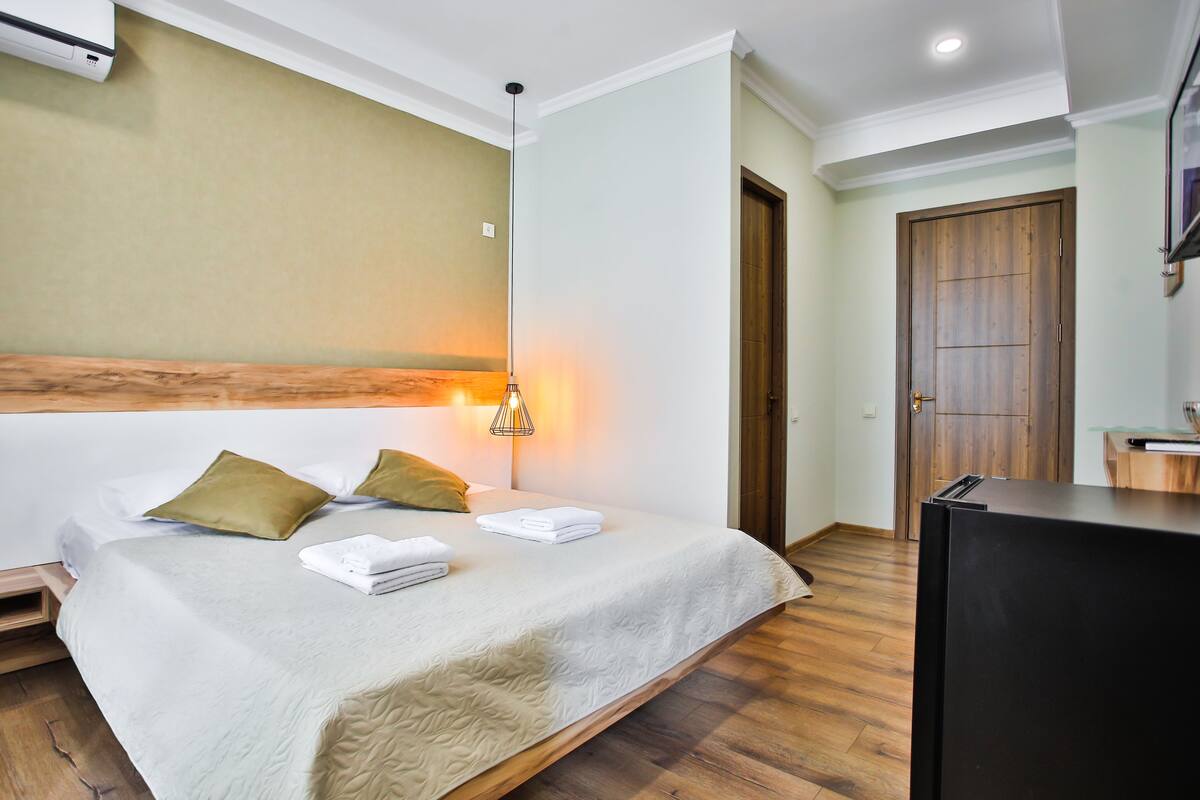 A hotel room with a bed, a wooden headboard, a wall-mounted air conditioner, a wooden door, and a black refrigerator.