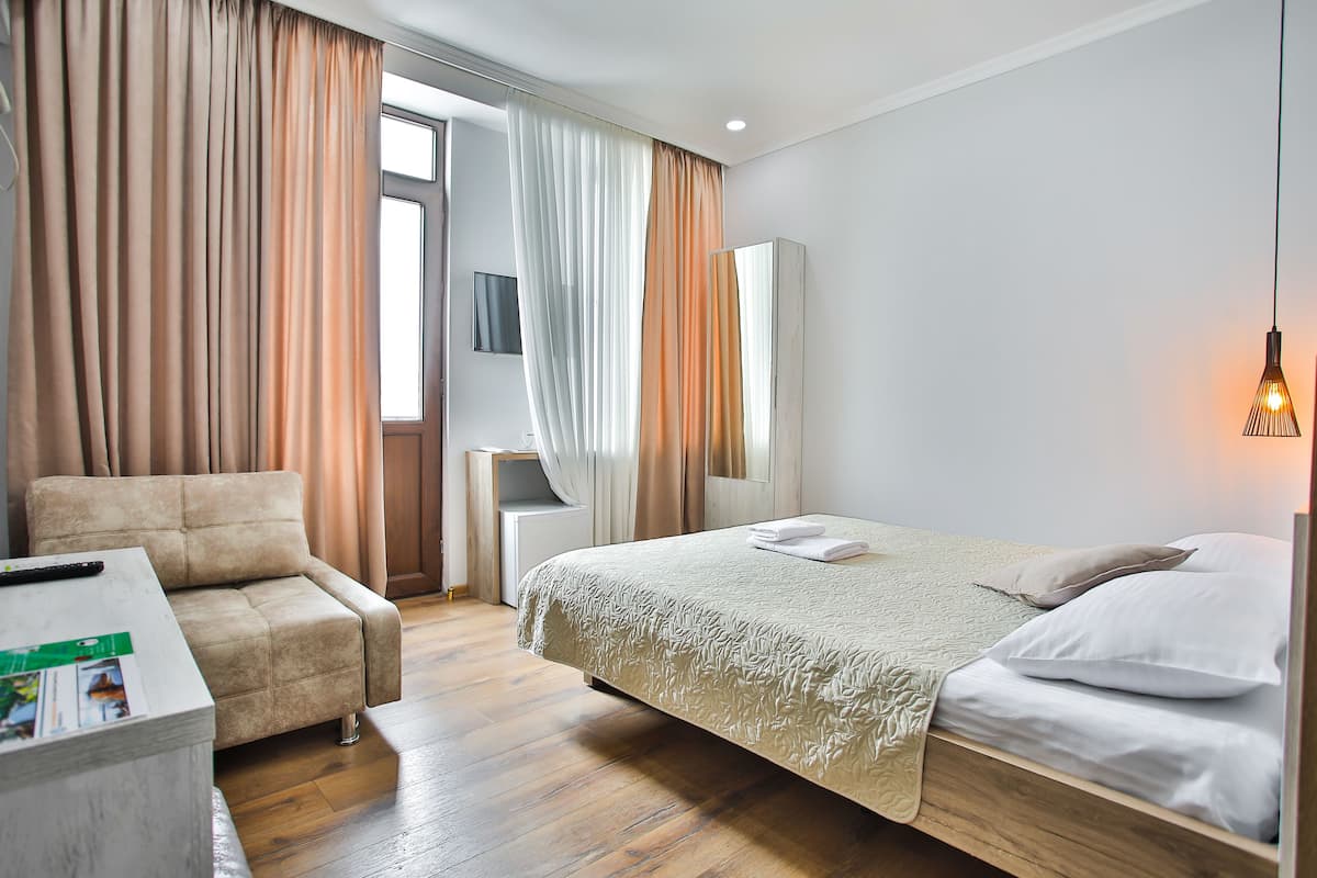 A hotel room with a sofa, a bed with white linens, a wooden floor, and a large window with curtains.
