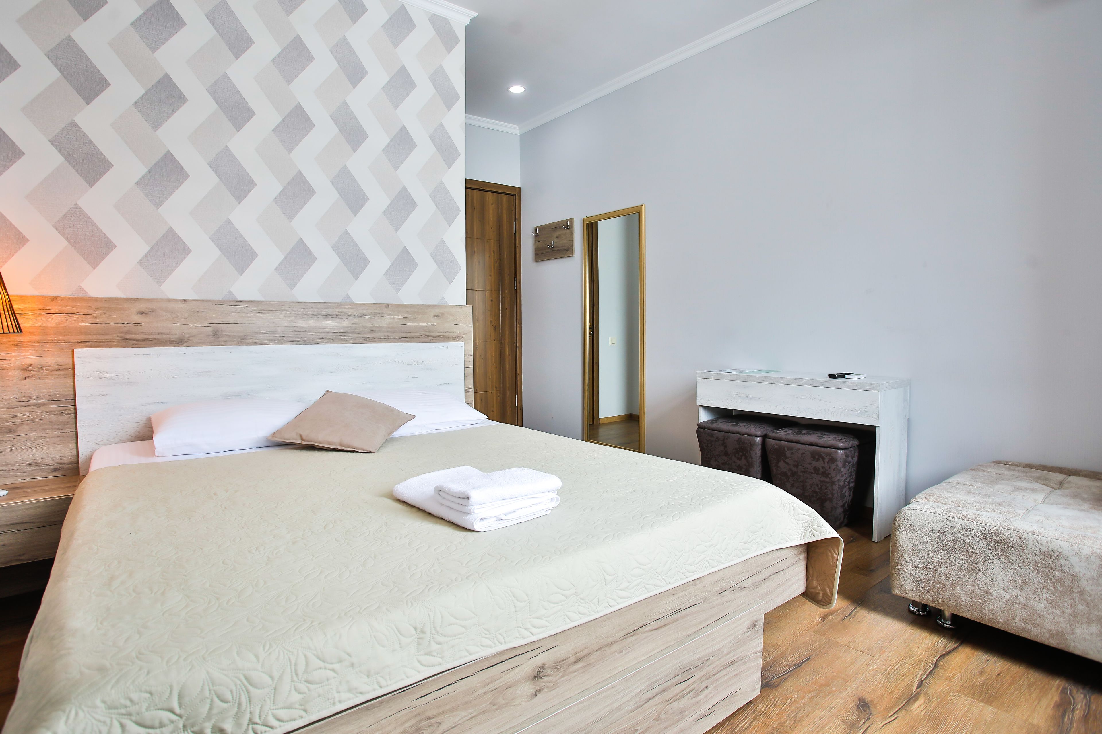 A neatly made bed with a white duvet and a folded towel, a wooden headboard, a wooden nightstand, a small desk with a mirror, and a bench.