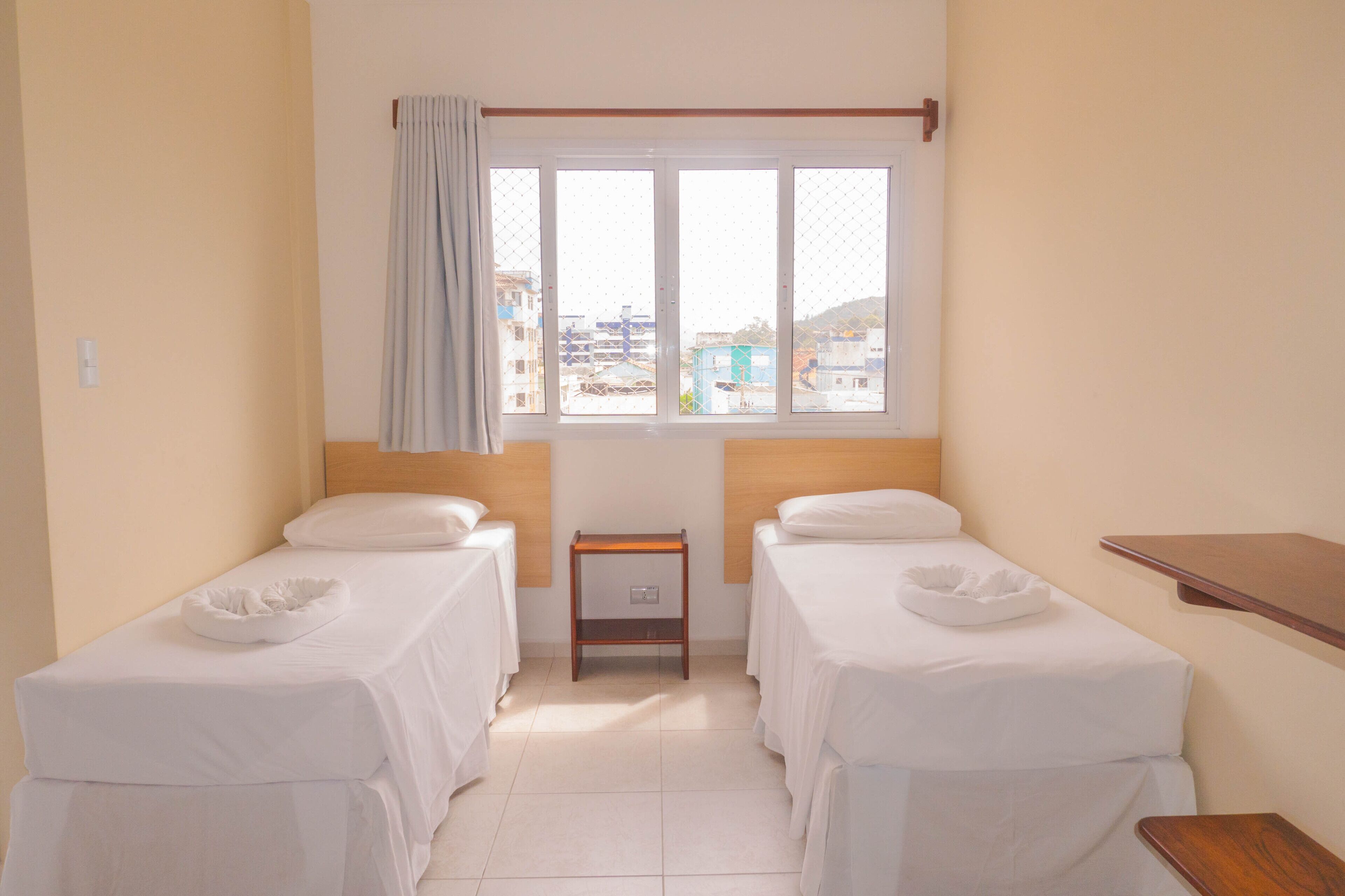 Ett hotellrum med två separata sängar, ett litet sängbord, ett fönster med utsikt över byggnader och en hylla i trä.