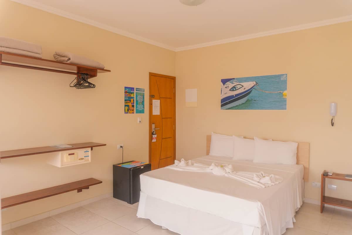 A hotel room with a bed, a wooden shelf, a painting of a boat, and a door.