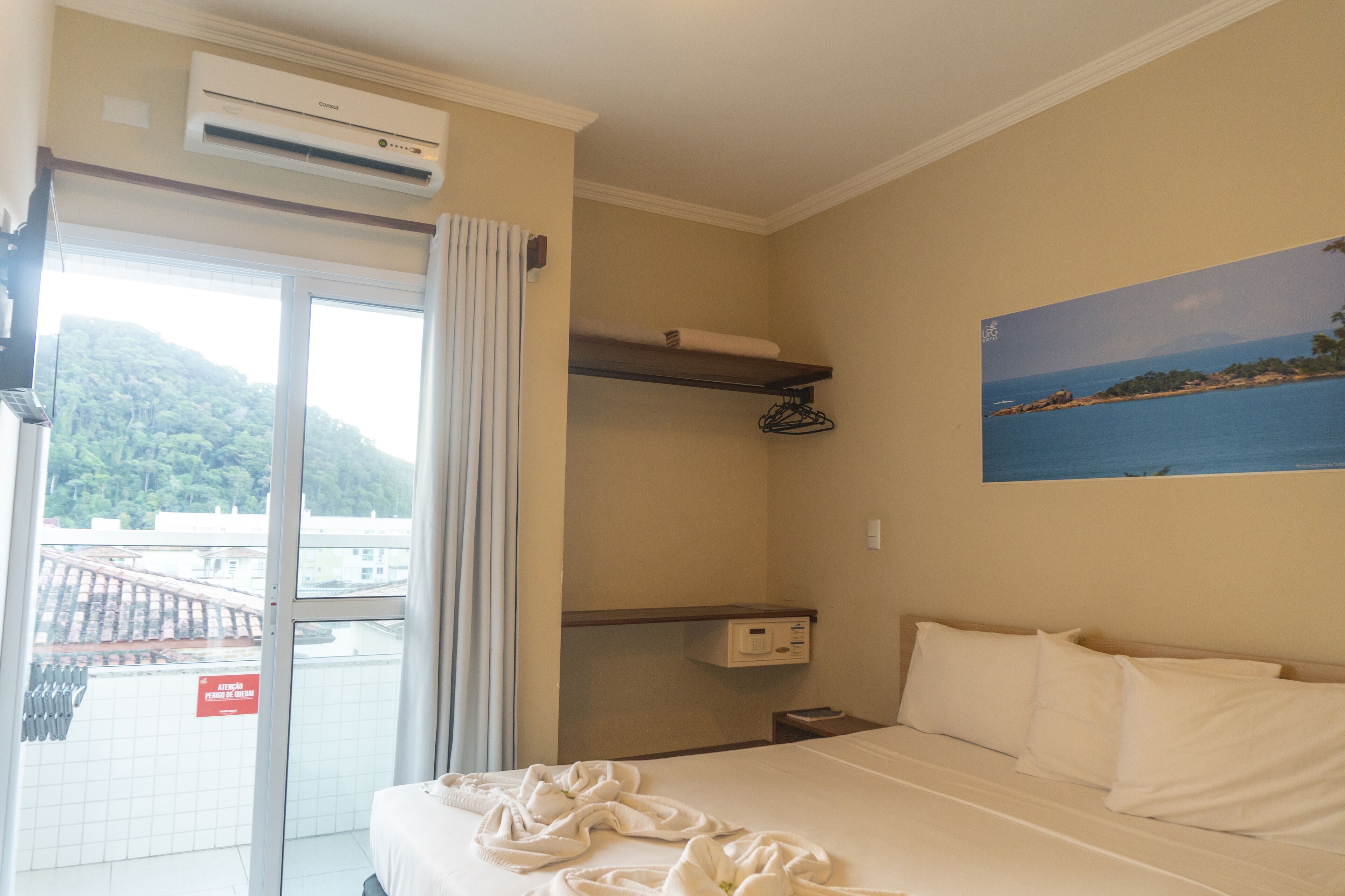 Ein Hotelzimmer mit einem Bett, einem Gemälde einer Küstenlandschaft, einem Fenster mit Blick auf eine Brücke und einer Klimaanlage.