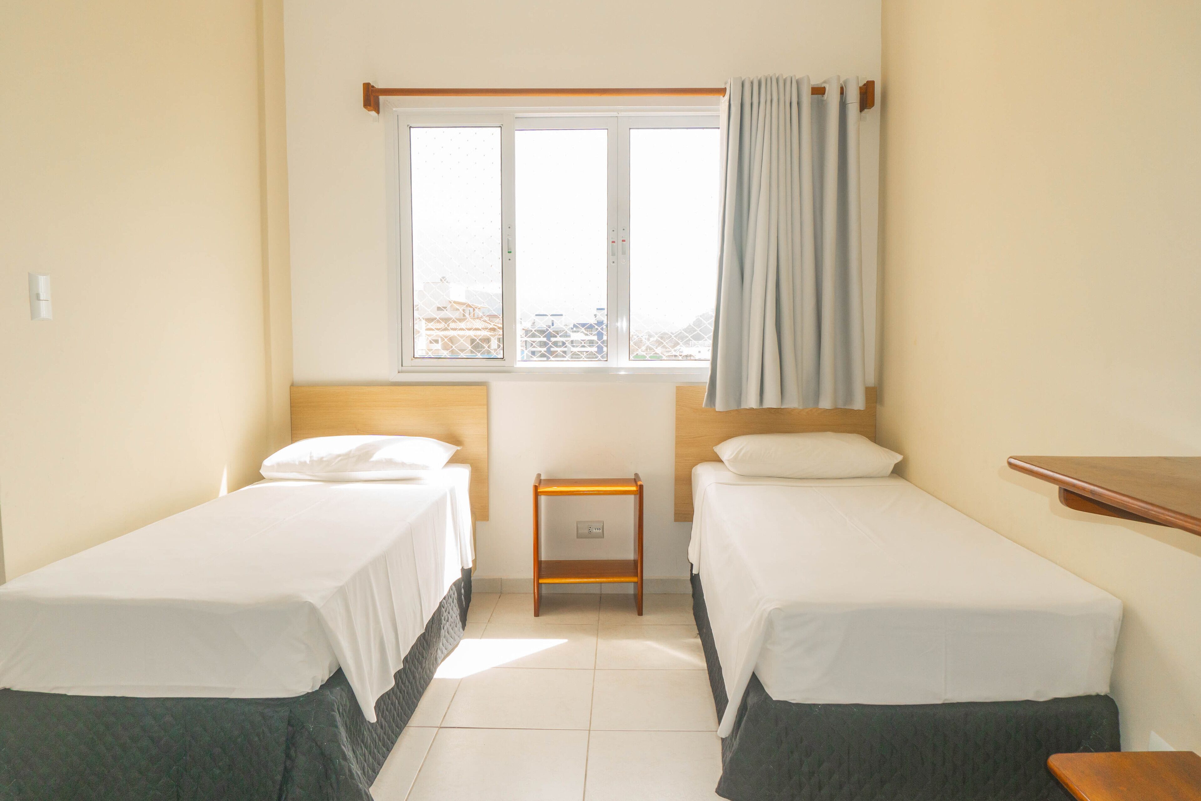 A hotel room with two single beds, a small wooden nightstand, a window with a view, and a curtain.