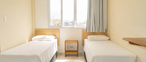 A hotel room with two single beds, a small wooden nightstand, a window with a view, and a curtain.