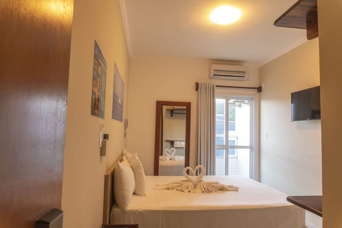 A hotel room with a bed, pillows, a flat-screen TV, an air conditioning unit, and a window with curtains.