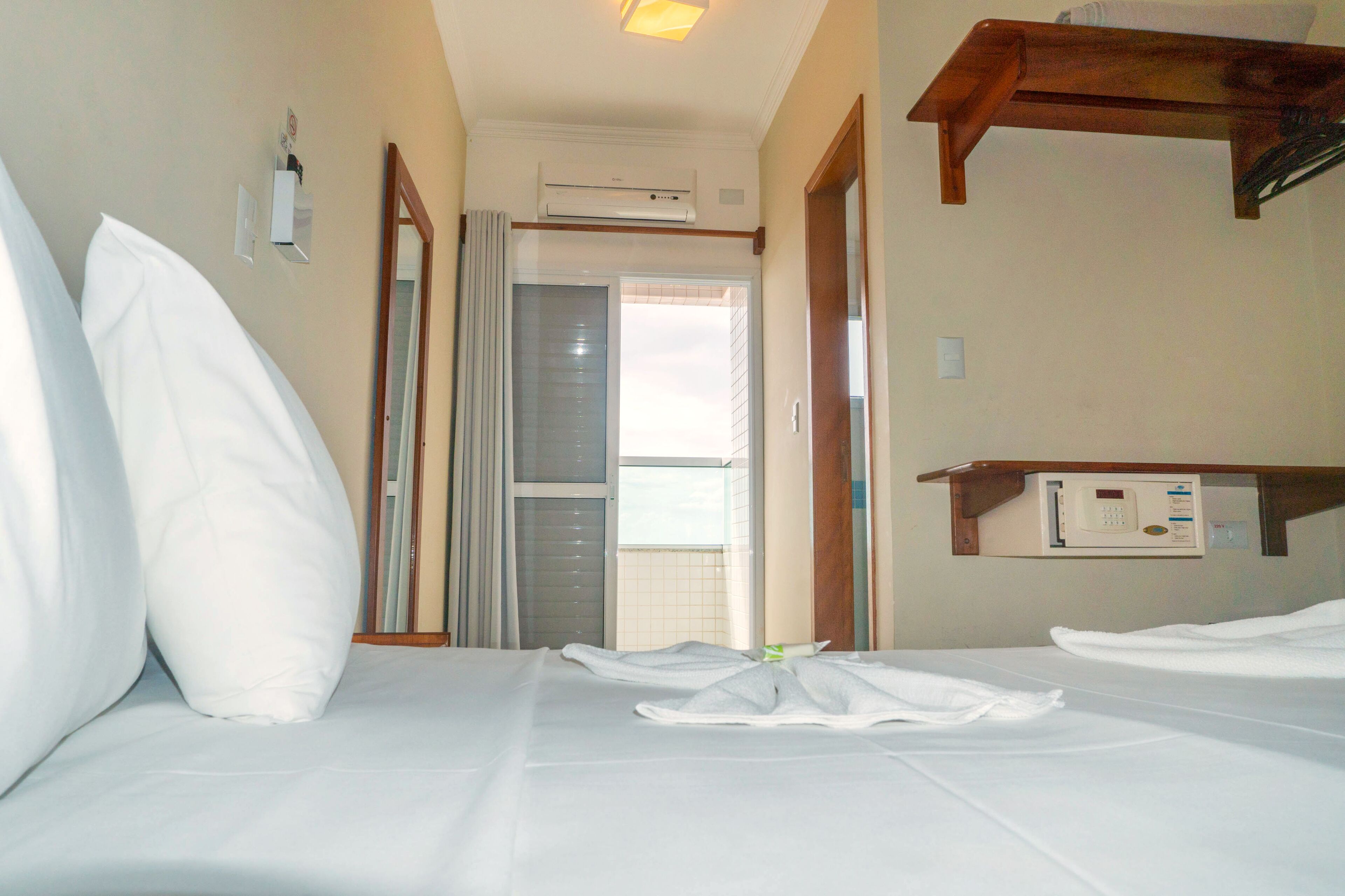 A hotel room with a bed, a bedside table, an air conditioning unit, and a window.