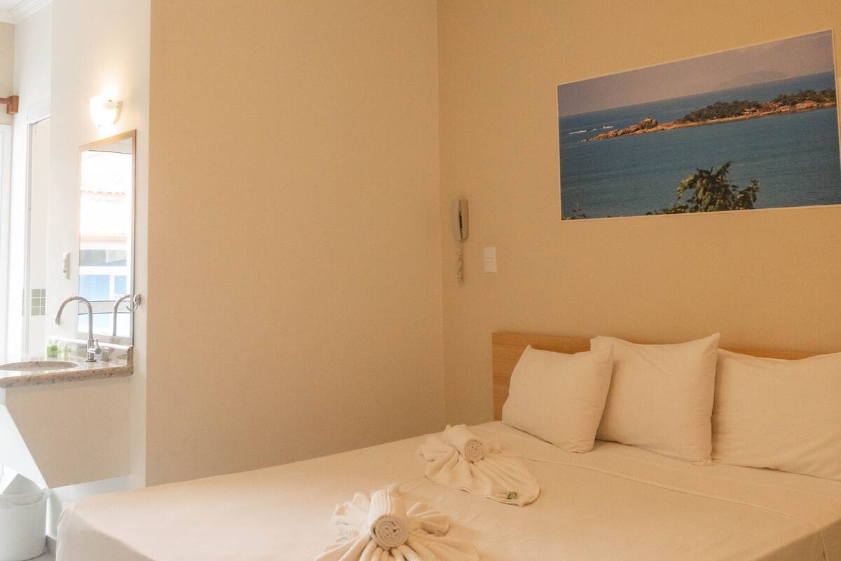 A hotel room with a bed, a painting of a beach, a bathroom with a sink and mirror, and a phone on the wall.