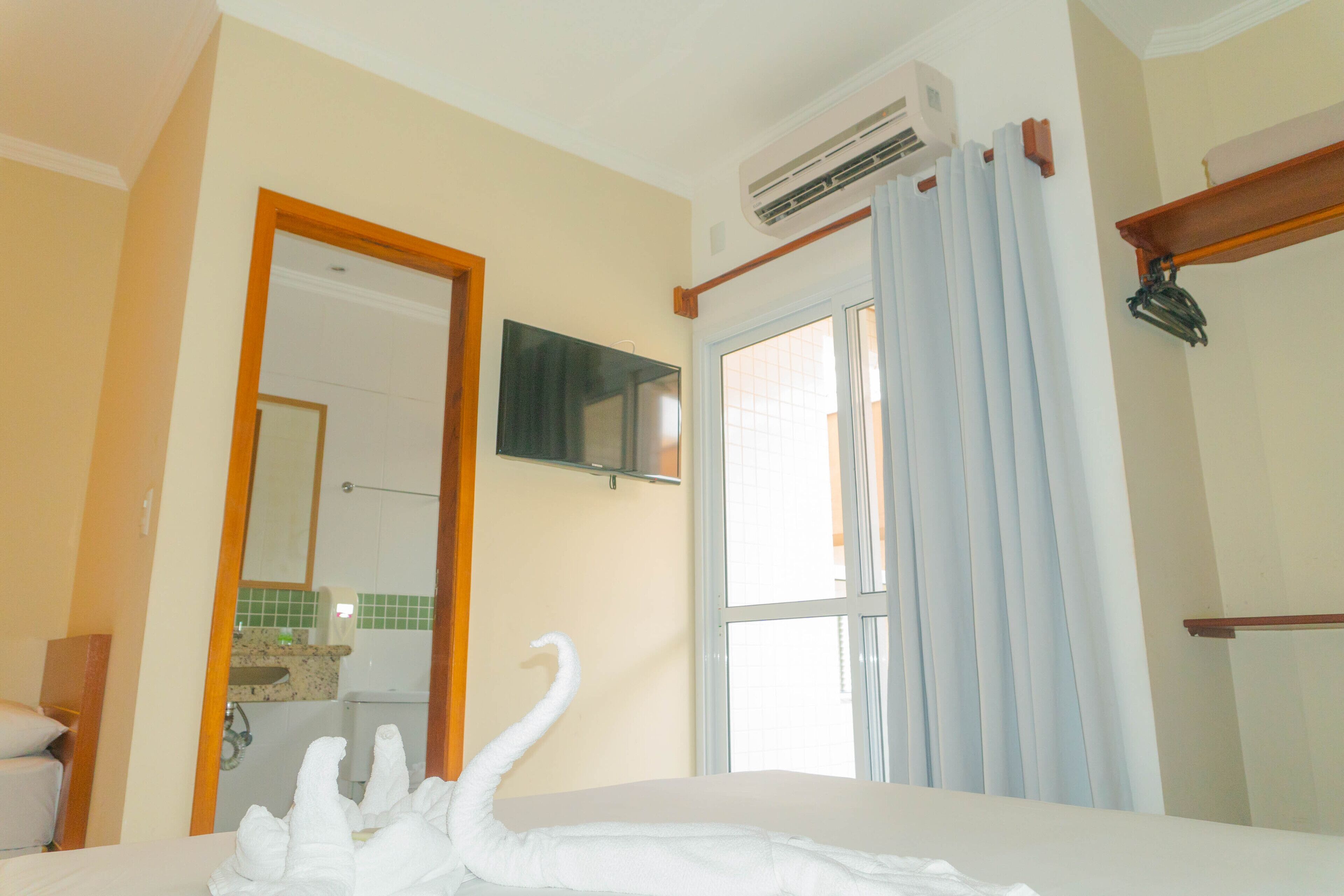 A hotel room with a bed, a bathroom visible through an open door, a mounted TV, and an air conditioning unit.