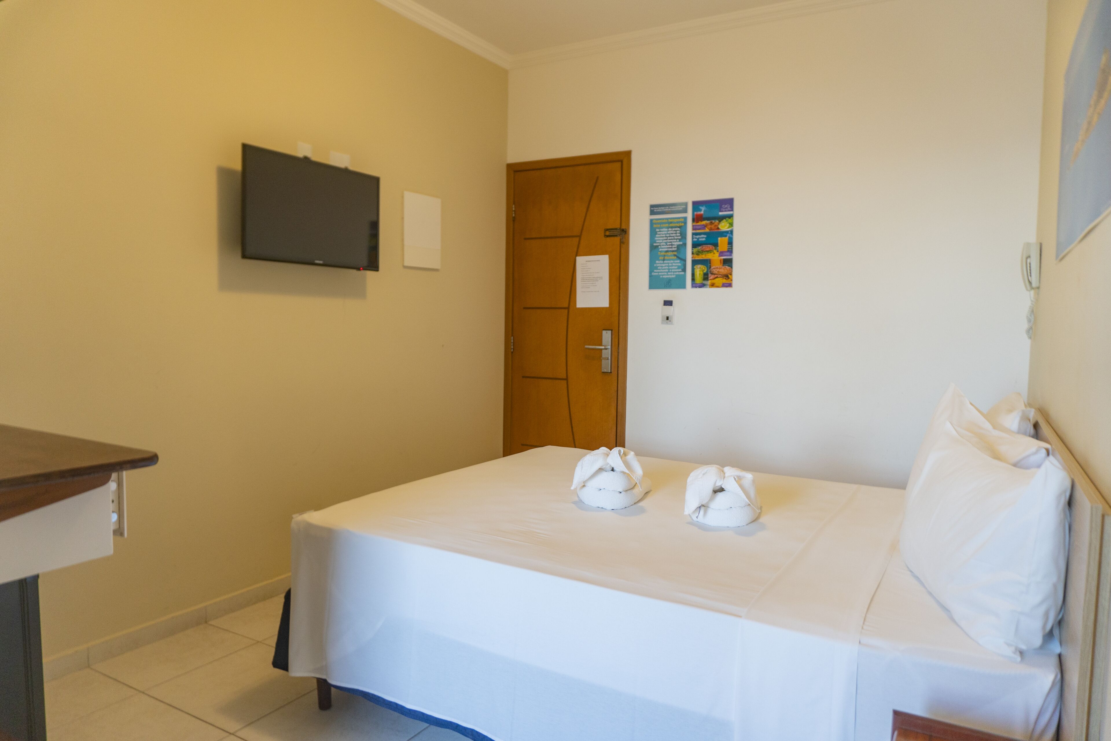 A hotel room with a bed, a television mounted on the wall, a wooden door, and a bulletin board with notices.