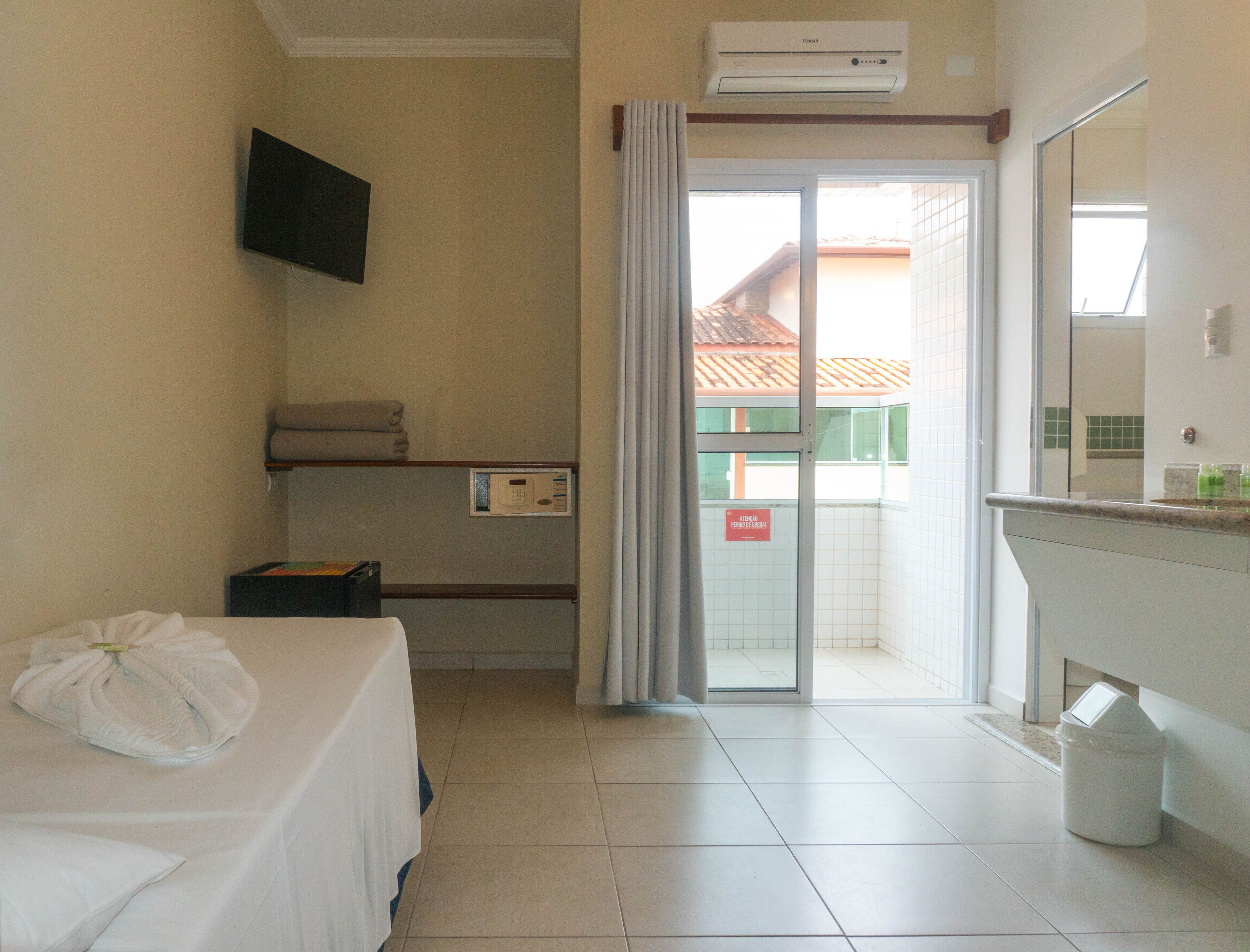 A hotel room with a bed, a television mounted on the wall, a bathroom with a shower, and a balcony with a view of buildings.