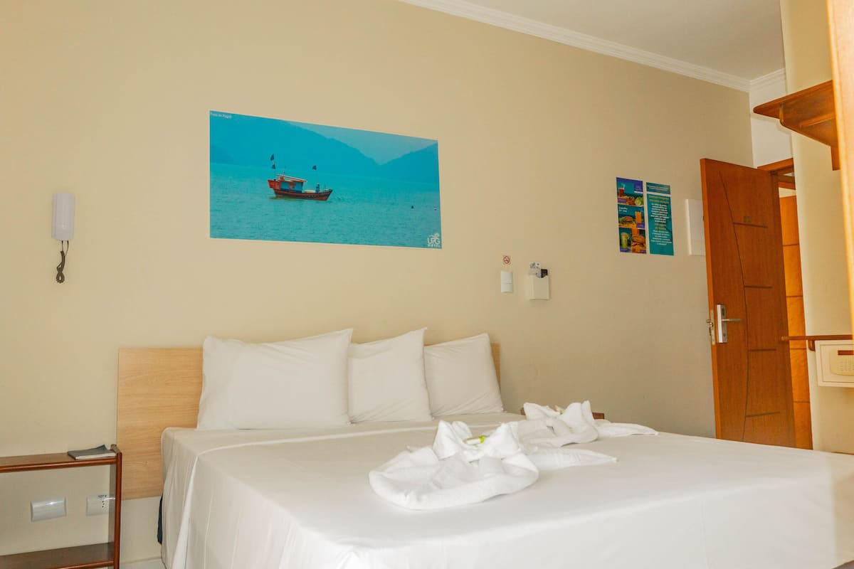 A hotel room with a bed, a painting of a boat on water, a wall-mounted light, and a door.