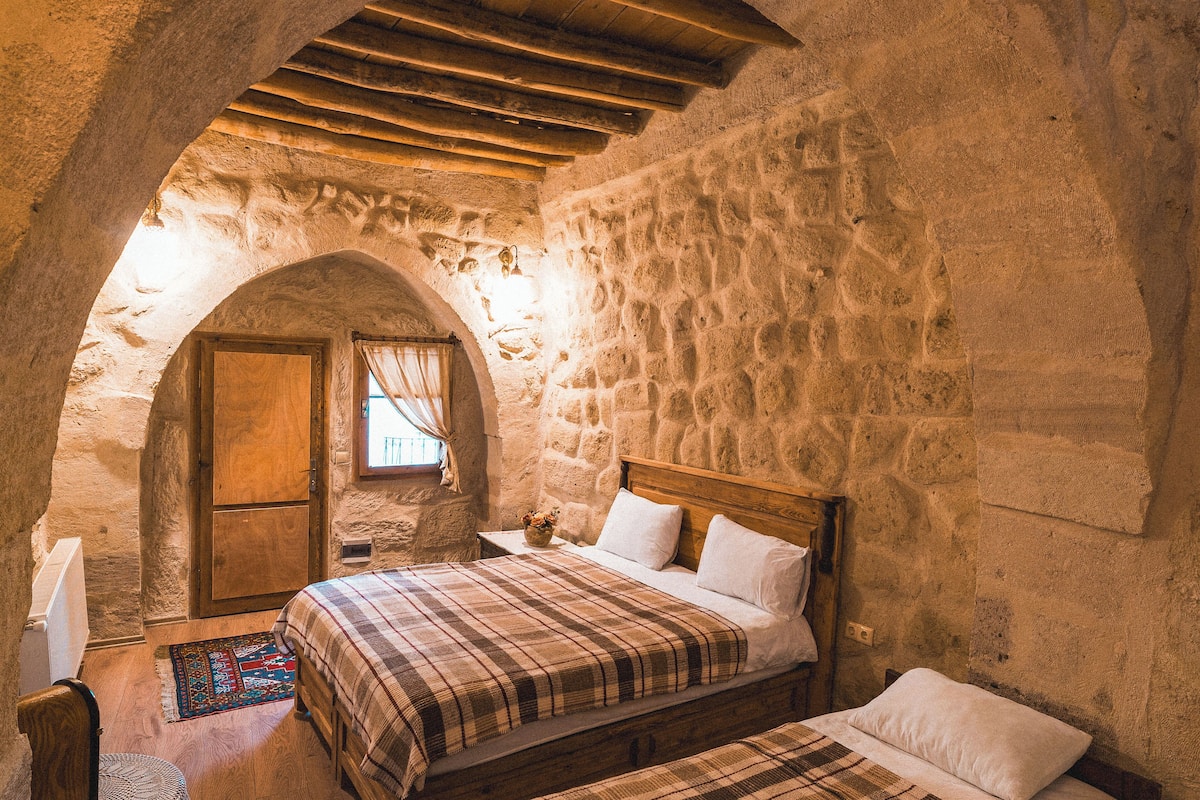 A rustic stone room with a wooden bed, a small table with a vase, and a window with curtains.