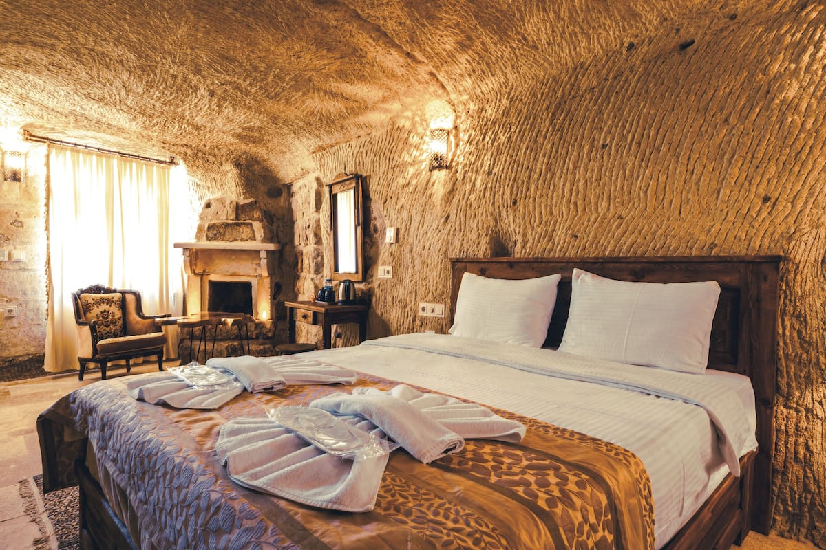 A rustic stone-walled bedroom with a wooden bed, a fireplace, and a stone bench.