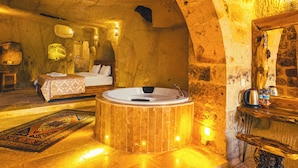 A luxurious room with a large wooden bathtub, a bed with white linens, and a stone wall. - Atillas Cave Hotel (Nevsehir)