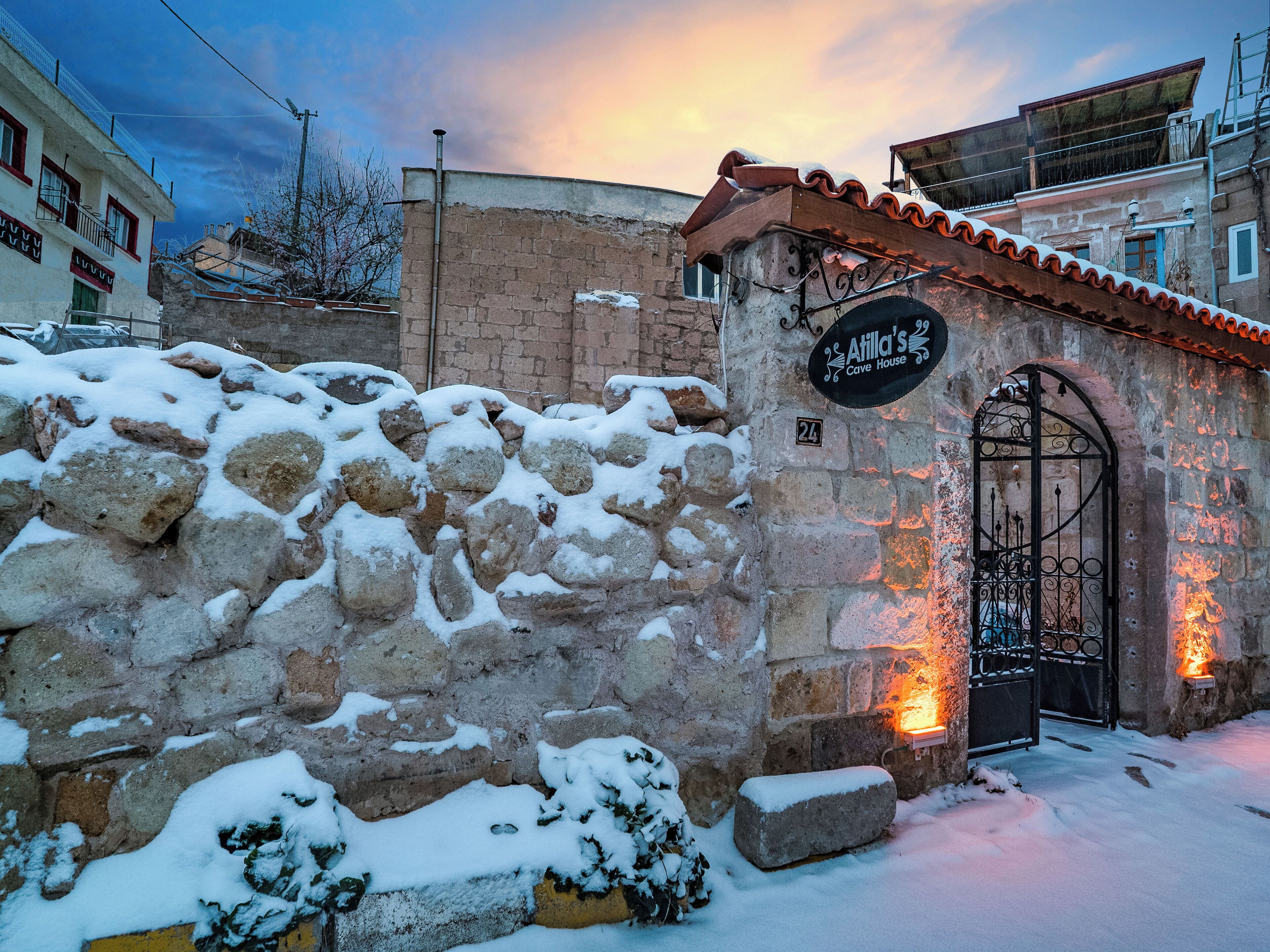 A stone wall with snow accumulation, a metal gate, and a building with a sign reading "Atilla's Cave House".