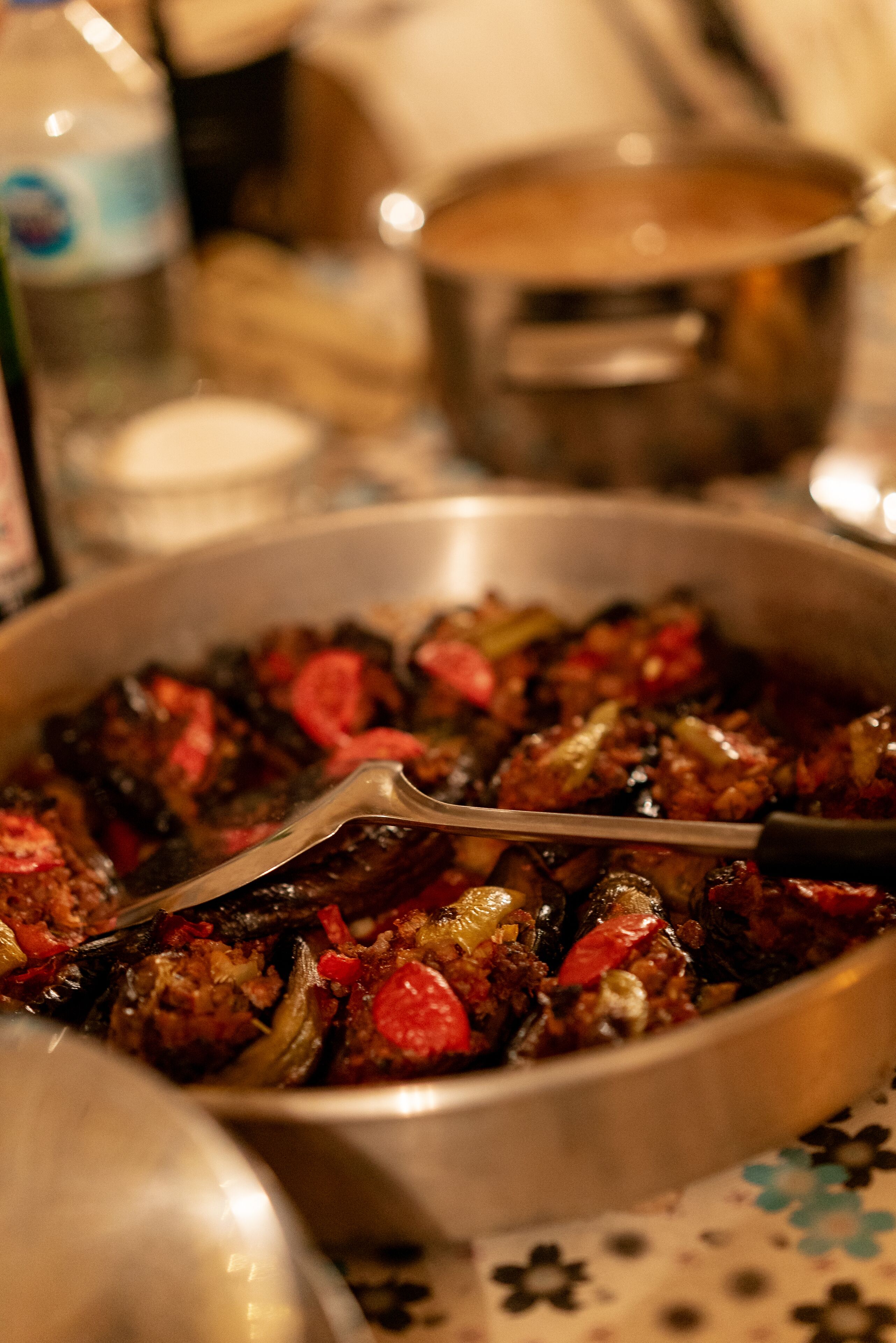 A close-up of a dish with meat and vegetables in a pan, with a bottle and a pot in the background.