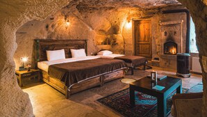A cozy, rustic bedroom with a stone ceiling, a fireplace, a wooden bed, and a small table with a rug. - Atillas Cave Hotel (Nevsehir)