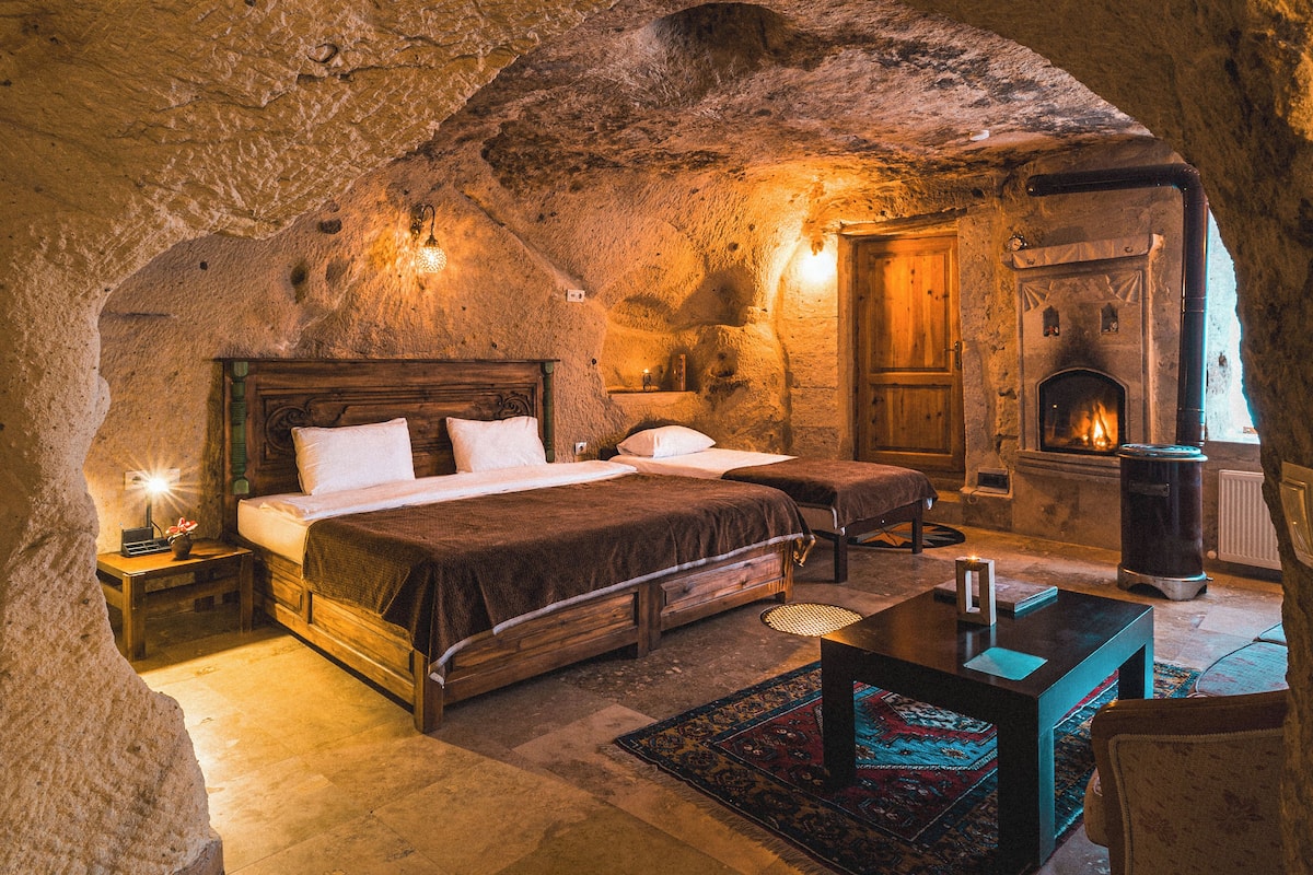 A cozy, rustic bedroom with a stone ceiling, a fireplace, a wooden bed, and a small table with a rug.