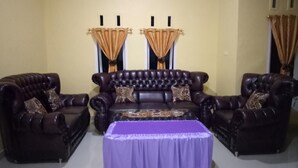 A living room with a dark brown leather sofa set, a matching armchair, and a coffee table with a purple tablecloth. - Sinar Harapan (Tomia Island)