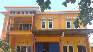 A two-story building with a yellow facade, a covered entrance, and potted plants in front. - Sinar Harapan (Tomia Island)