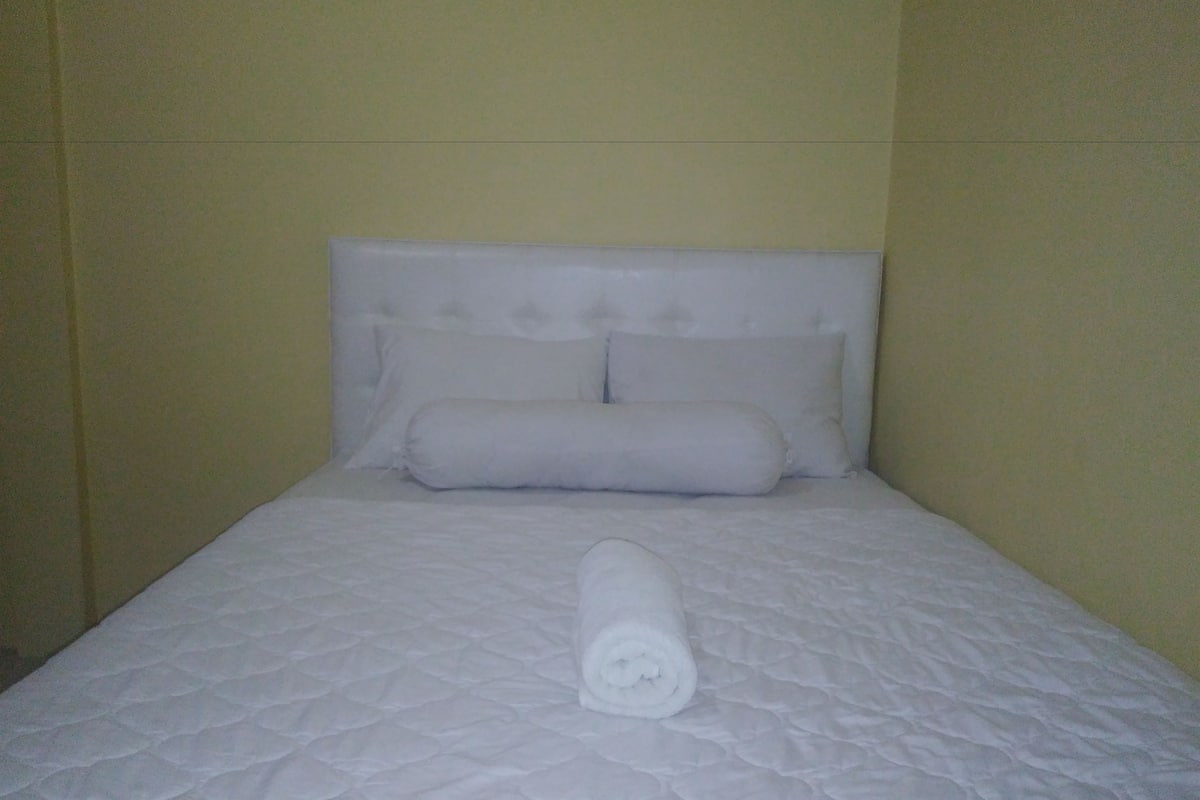 A neatly made bed with a quilted cover, pillows, and a rolled towel.